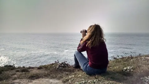 female student with curly hair is sitting on a bluff and looking out at the ocean through binoculars