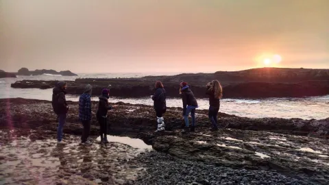 six students stand in the rocky intertidal zone looking out at the sunset over the Pacific Ocean